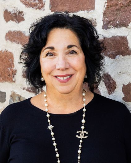 Woman with short dark hair smiles, wearing a black cardigan and beaded necklaces, leaning against a green wall.
