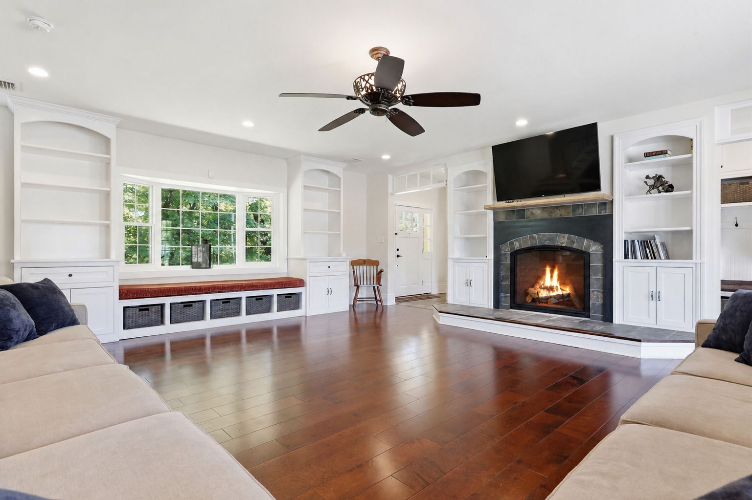 Bright living room with beige sofas, hardwood floors, fireplace, built-in shelves, and large window seat.