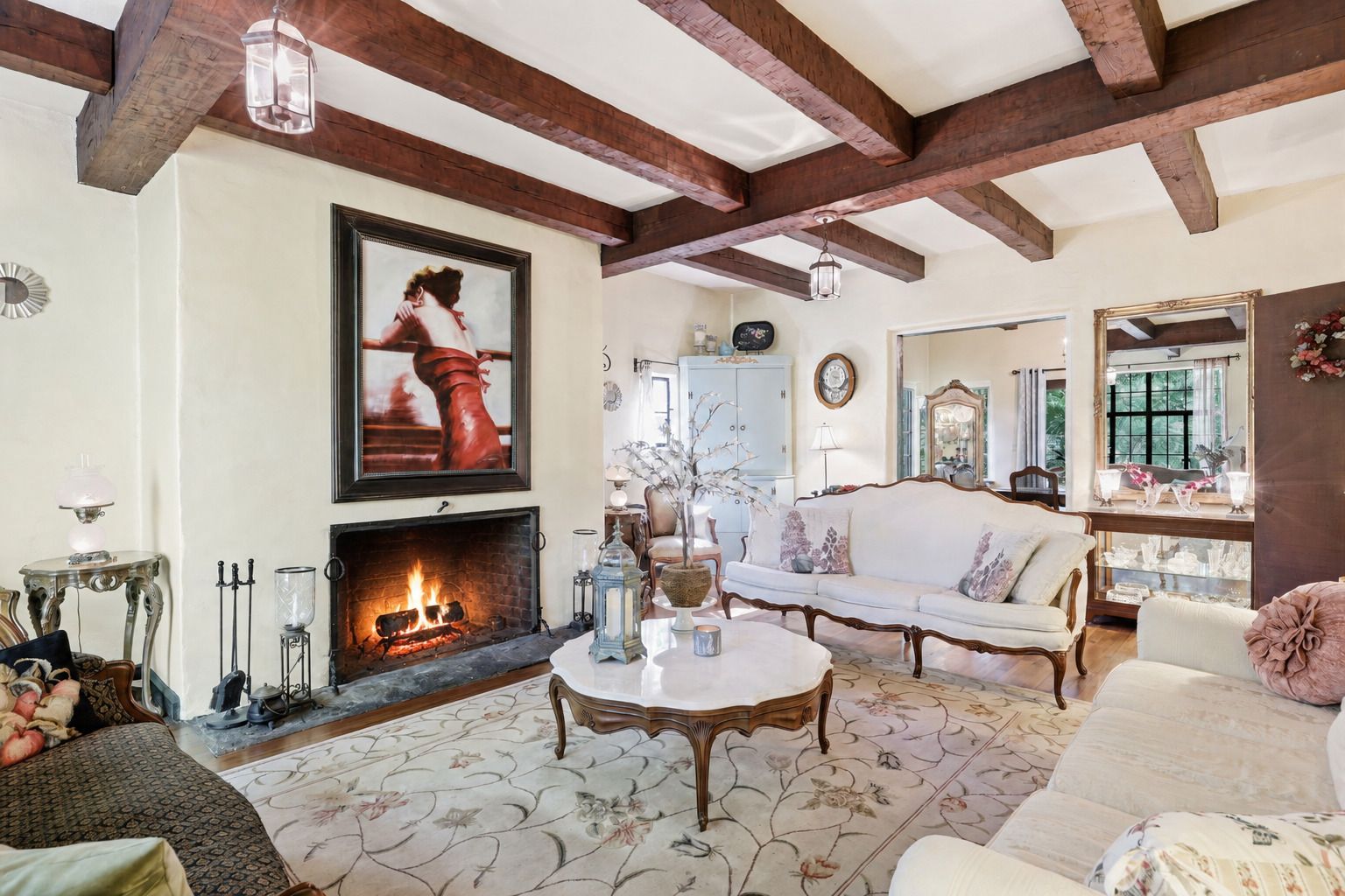Cozy living room with exposed beams, a lit fireplace, beige sofa, and framed art above the mantel