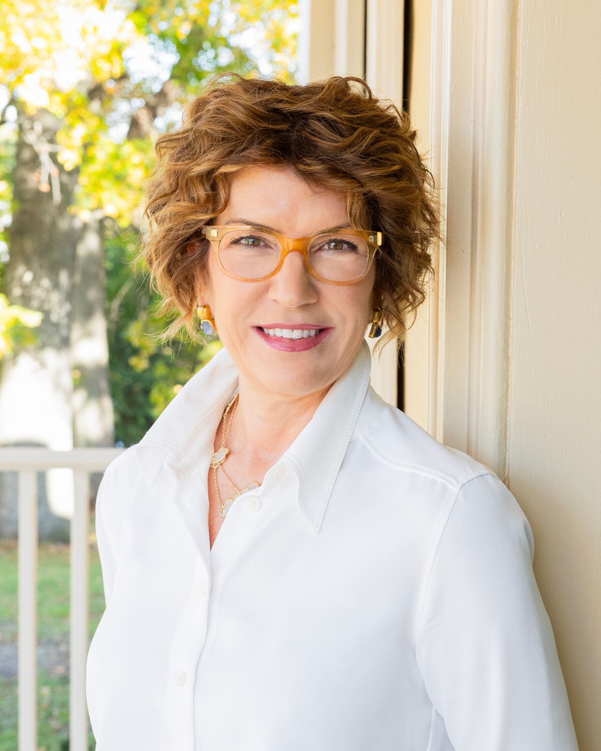 A person with curly hair and glasses, wearing a white collared shirt, smiles while leaning against a wall outdoors.