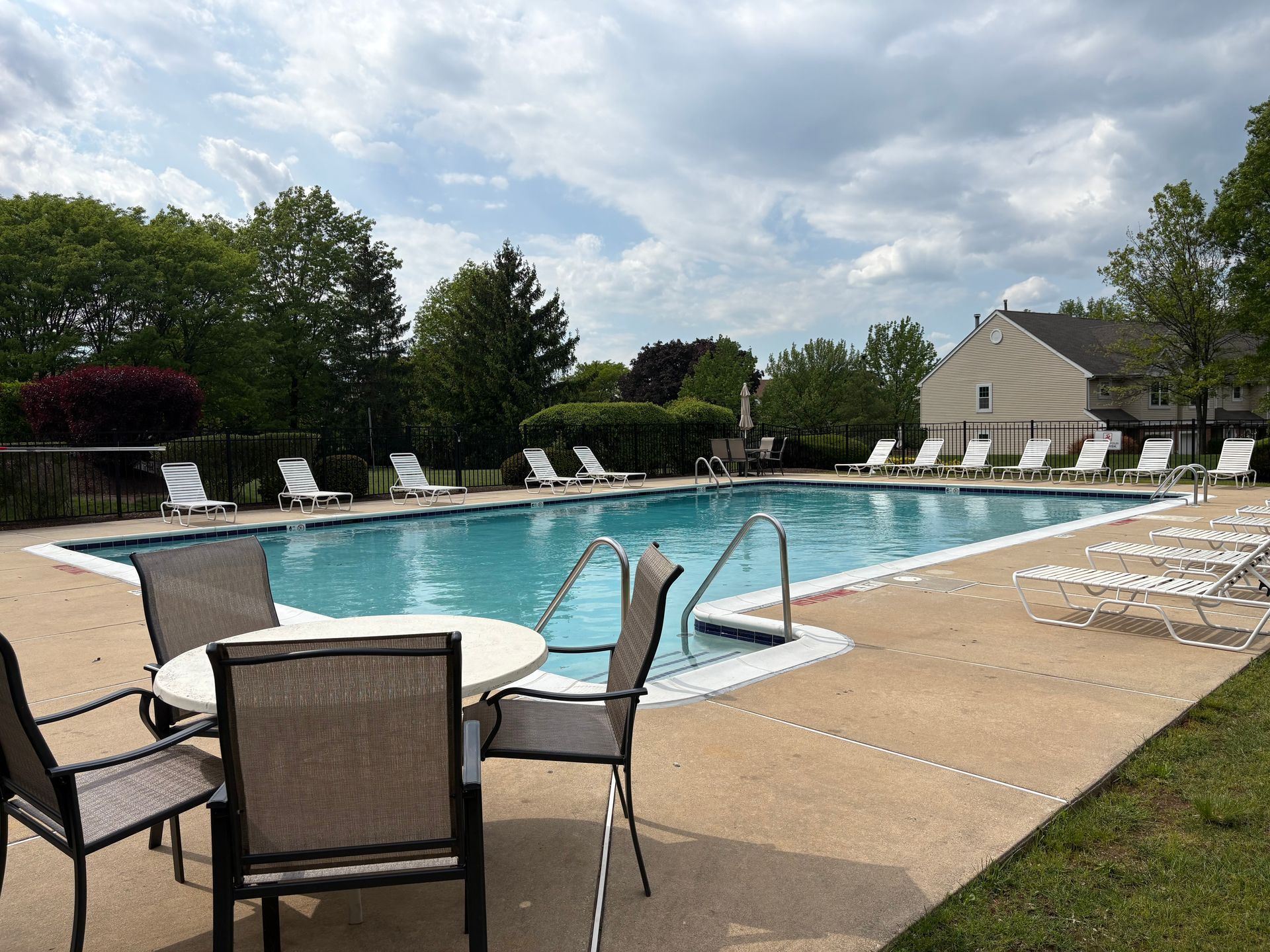 Outdoor swimming pool with lounge chairs, patio tables, and blue water under a cloudy sky