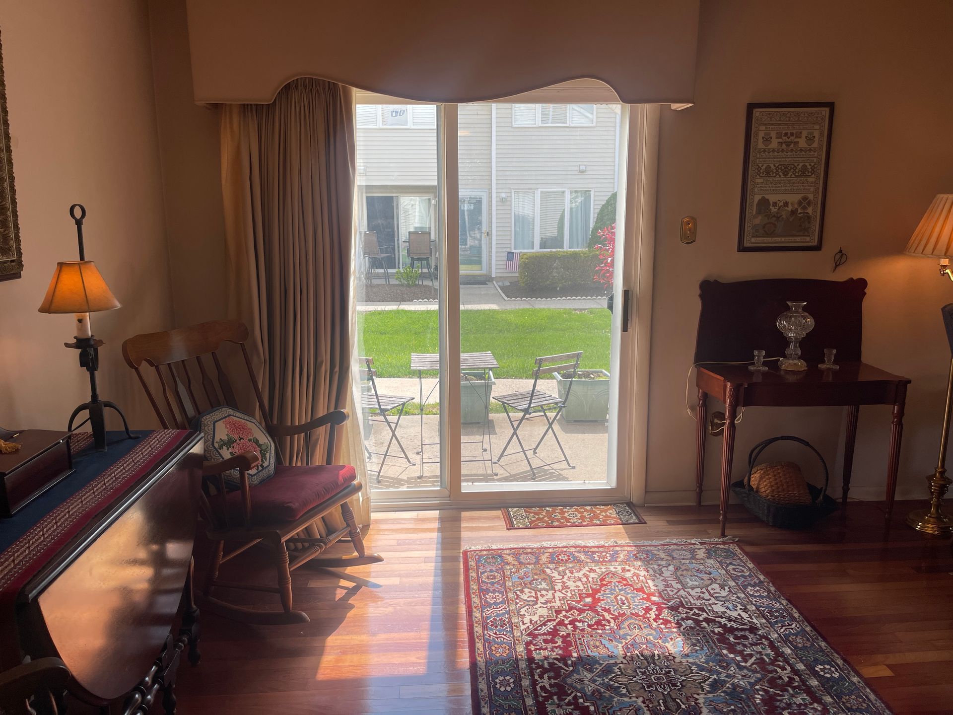 Sunlit living room with sliding glass door, patterned rug, lamps, and patio view beyond curtains