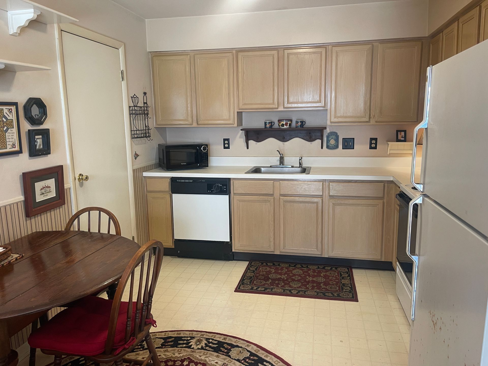 Small kitchen with light wood cabinets, white appliances, dining table, and patterned rug.