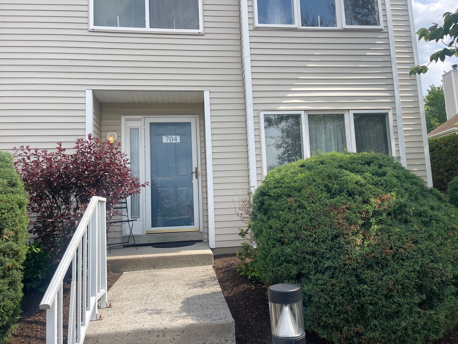 Front entrance of a beige townhouse with a glass door, steps, and trimmed bushes along a walkway