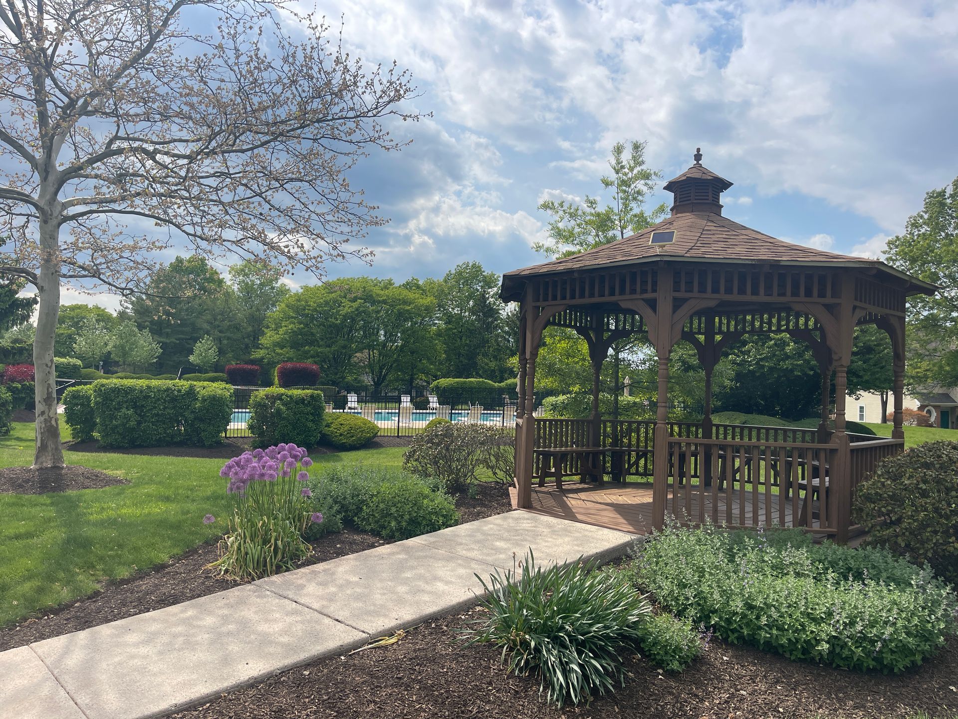 Garden gazebo beside a path, surrounded by flowers and greenery under a partly cloudy sky