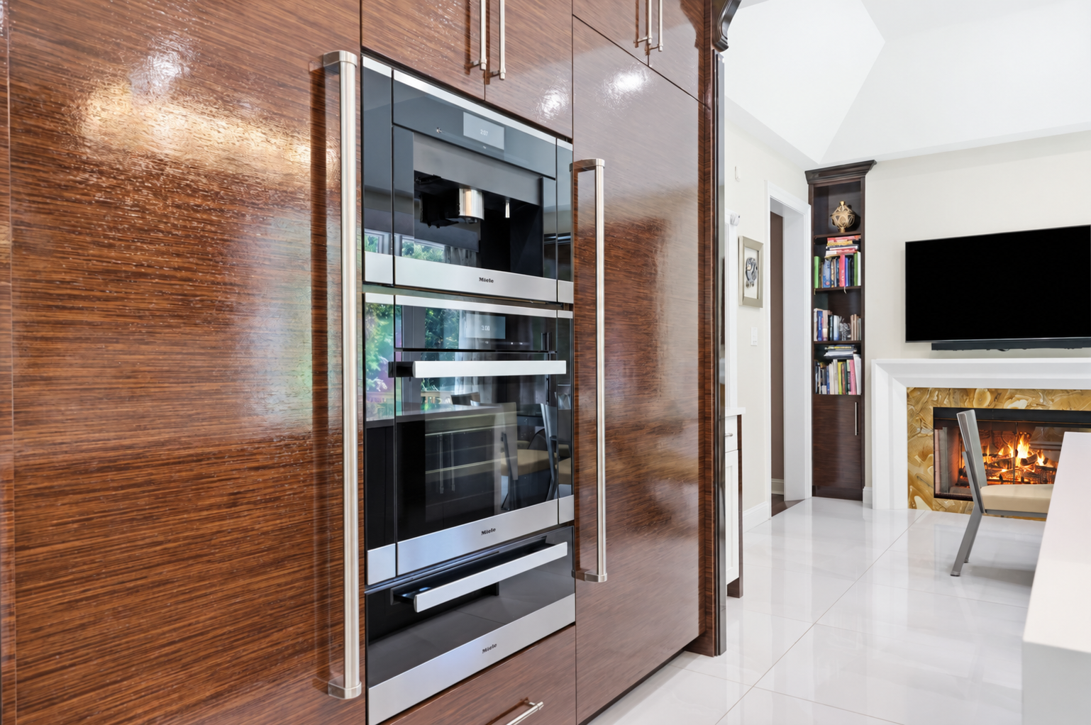 Modern kitchen with glossy wood cabinets, built-in oven, and bright living area with TV and fireplace beyond