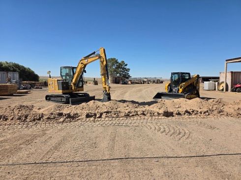 Excavator and skid steer on a construction site with dirt piles under a clear blue sky