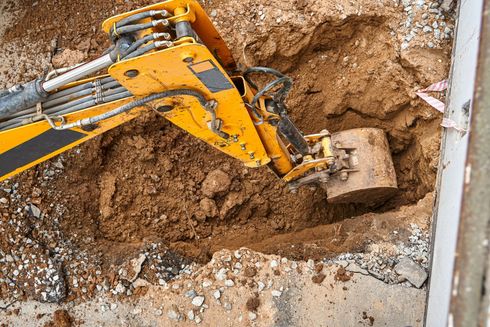 Yellow excavator digging soil at a construction site, with a bucket scooping dirt near a trench