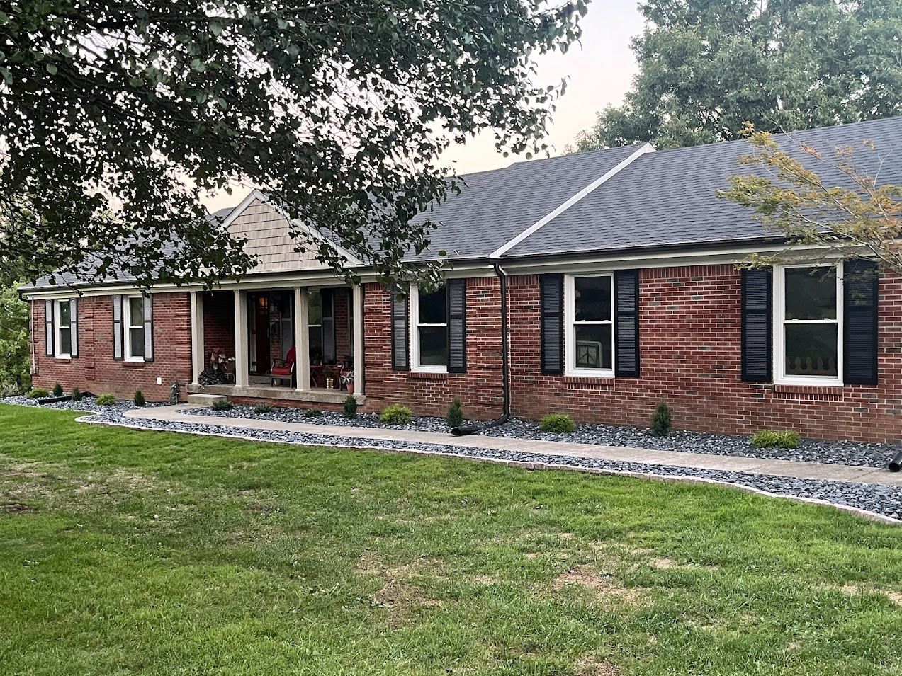 Brick house with black shutters, white-trimmed windows, and front porch, under a tree on a green lawn.