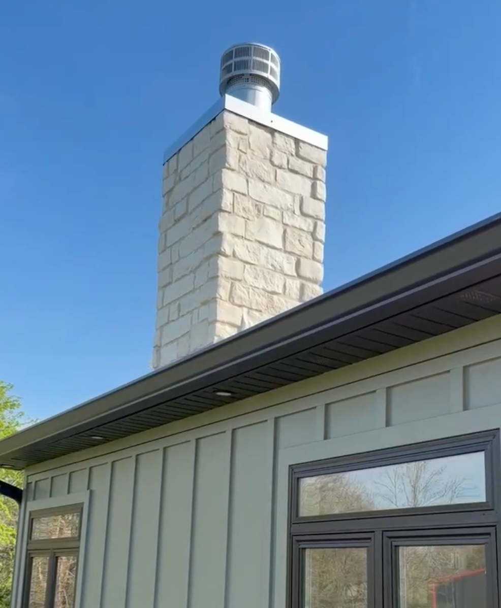 Stone chimney with a metal cap above a light gray house under a blue sky