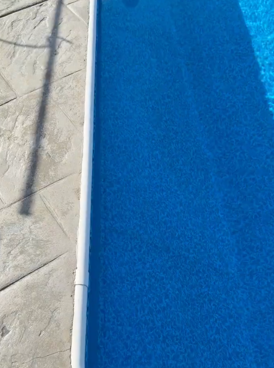 Blue swimming pool beside a white tiled deck with a shadow across the pavement.