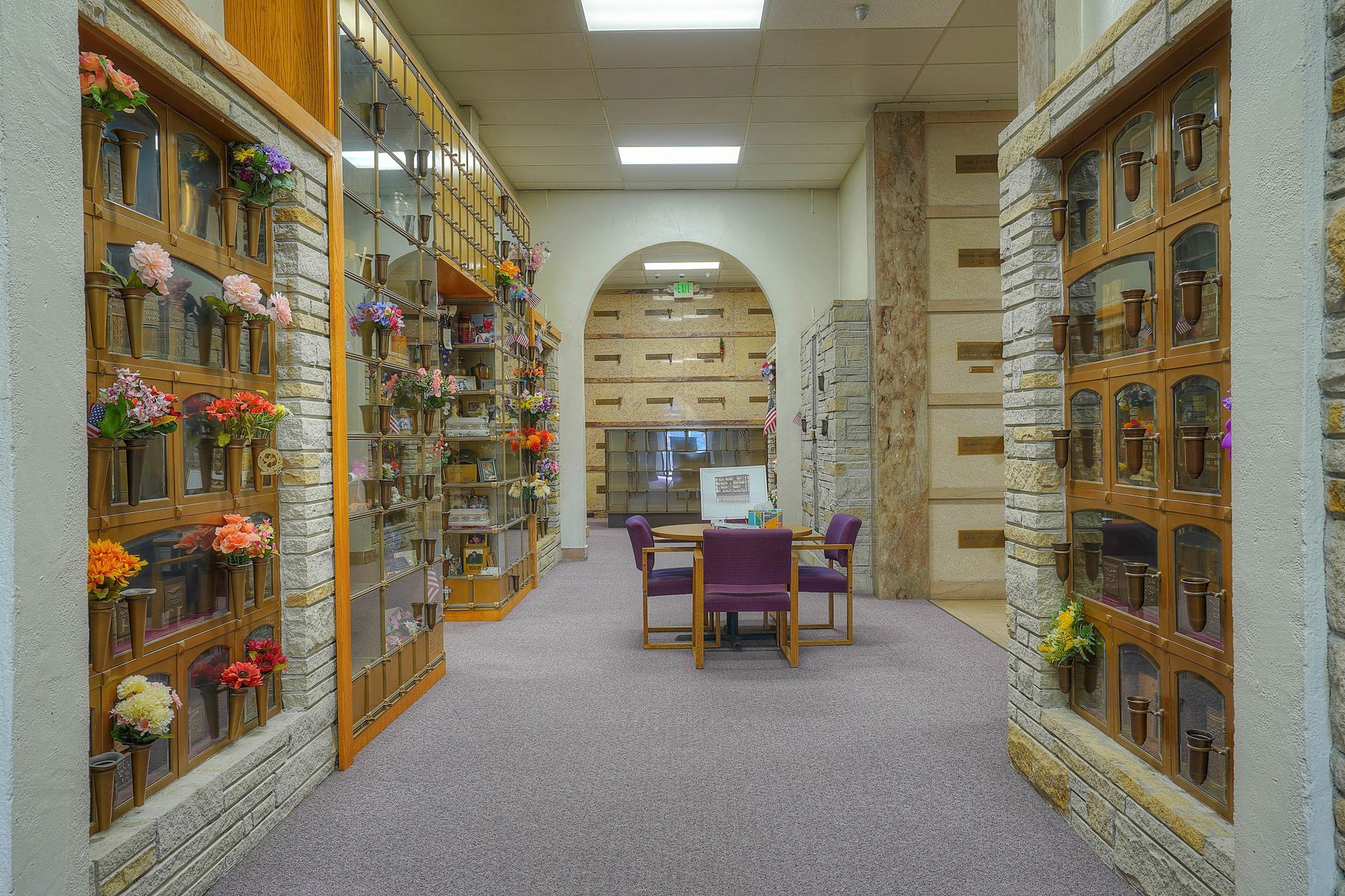 Hallway with wall-mounted memorial niches and flowers, leading to a small seating area with purple chairs