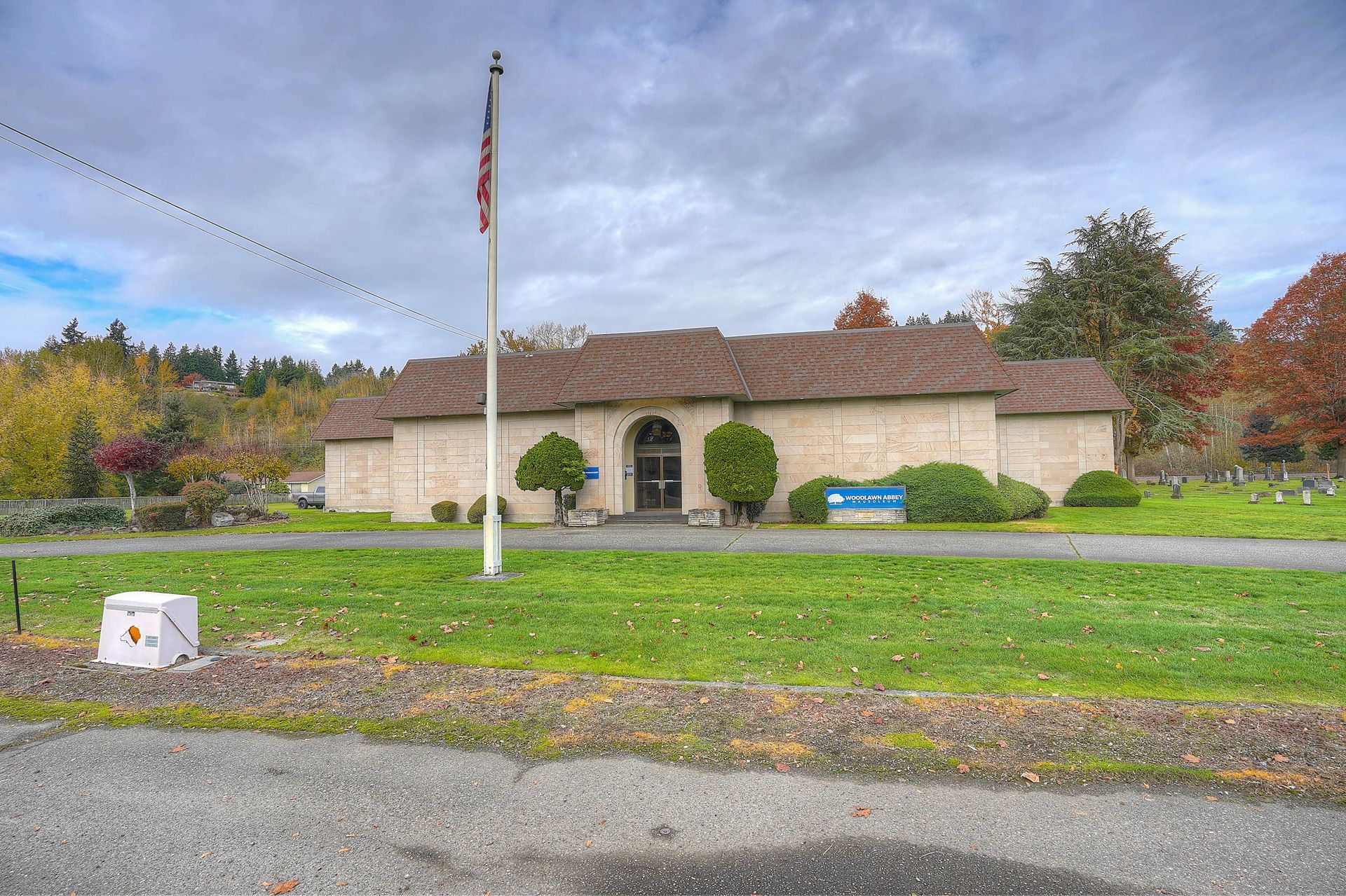 Single-story beige building with a flagpole, blue bench, and landscaped lawn under a cloudy sky