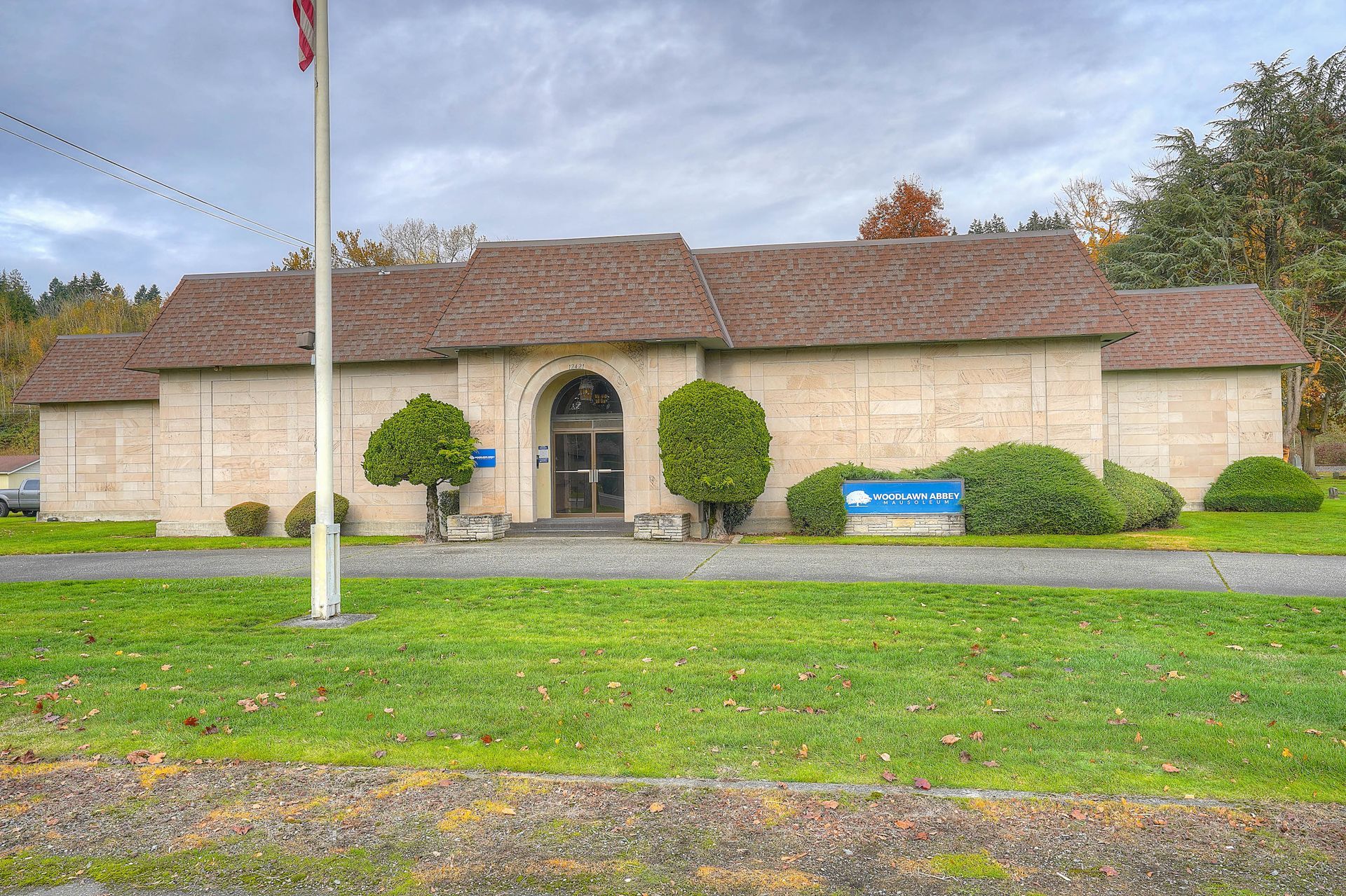 Single-story beige building with arched entrance, trimmed shrubs, and a blue sign beside a grassy roadside.