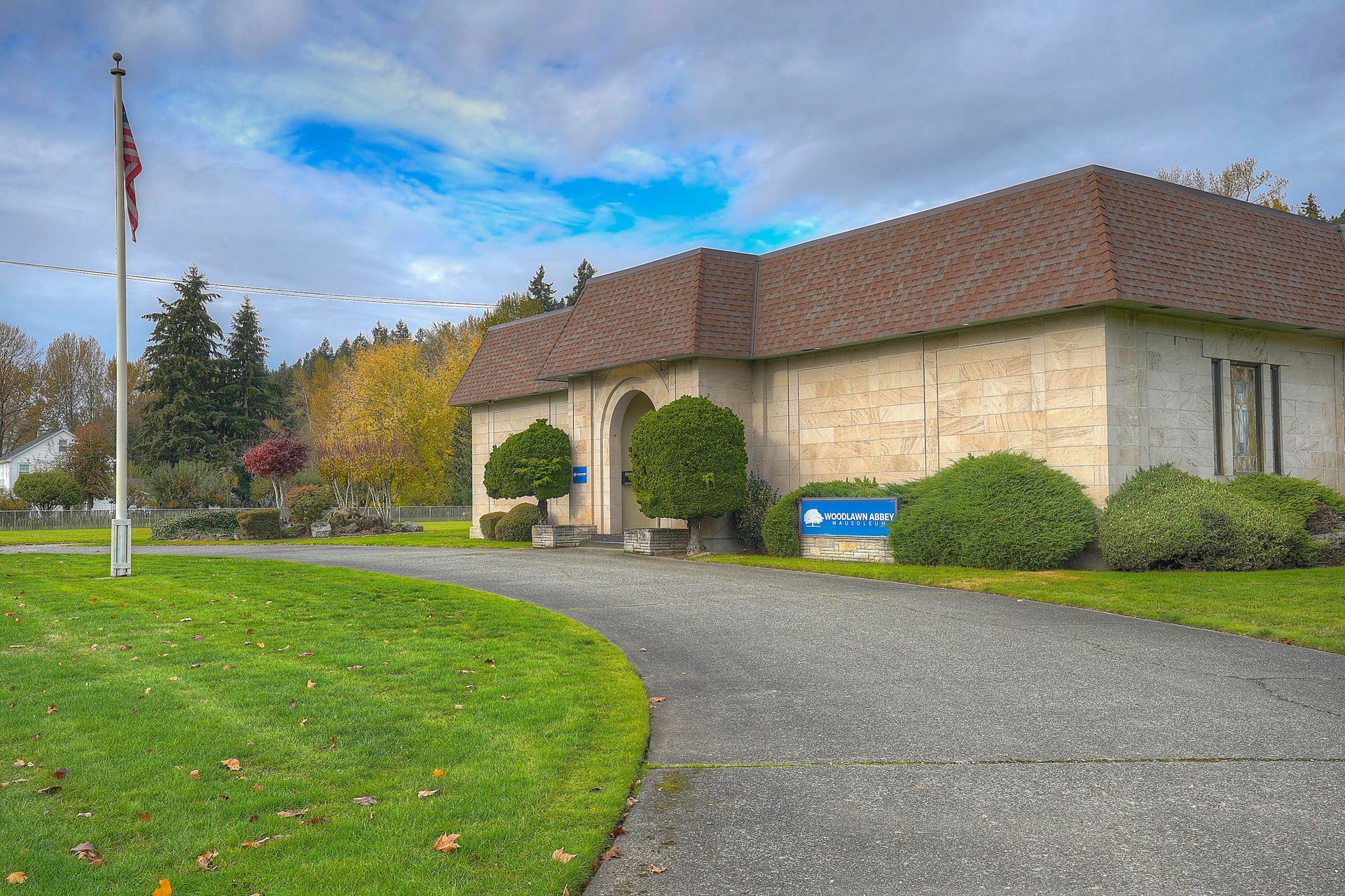 Stone building with brown roof beside a gravel driveway, green lawn, and flagpole under a cloudy sky