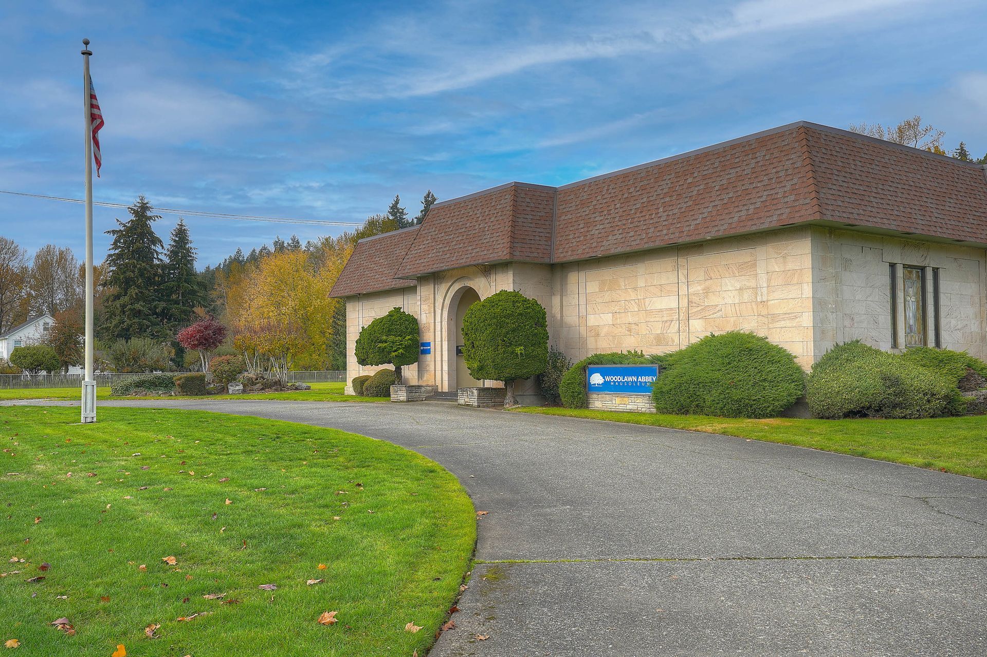 Stone building with brown roof, gravel driveway, lawn, and a flagpole under a cloudy sky