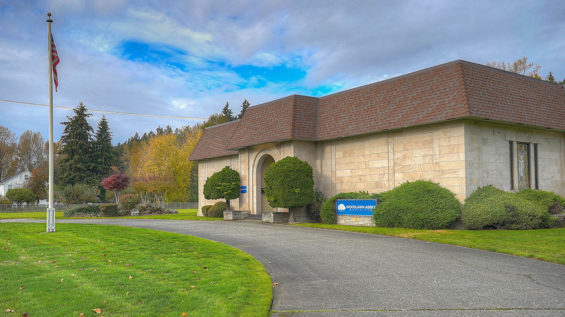 Stone building with brown roof, blue sign, and curved gravel path beside green lawn and trees