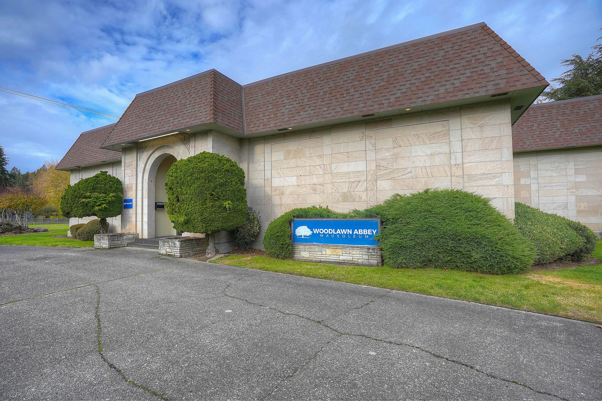 Stone building with brown roof, arched entrance, and blue sign beside a gravel driveway and shrubs