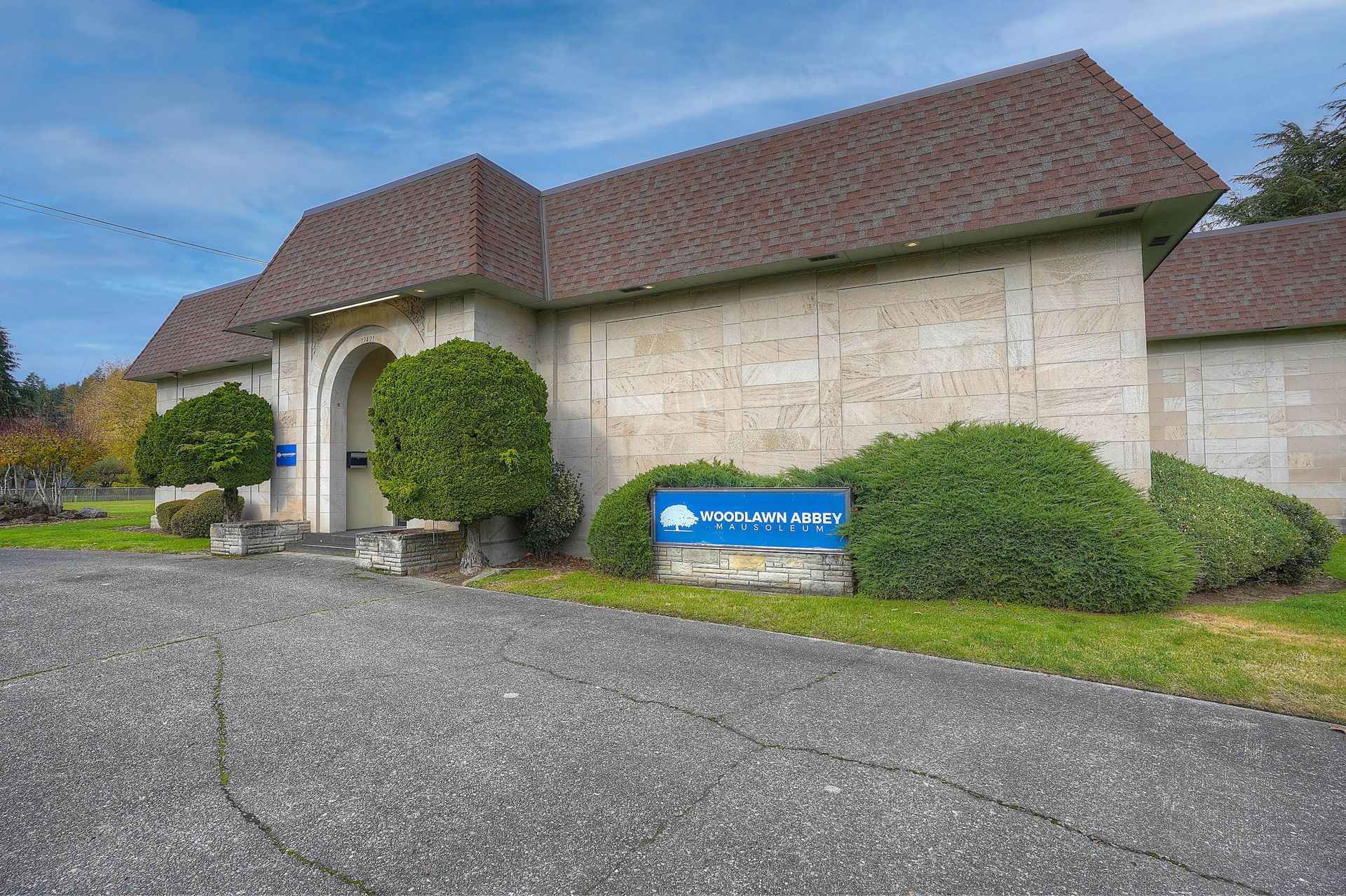 Stone building with brown tiled roof, trimmed hedges, and a blue sign beside a gravel drive.