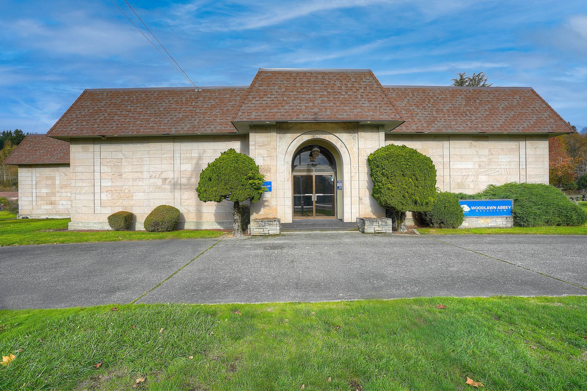 Stone building with a red-tiled roof, arched doorway, and trimmed shrubs beside a gravel drive.