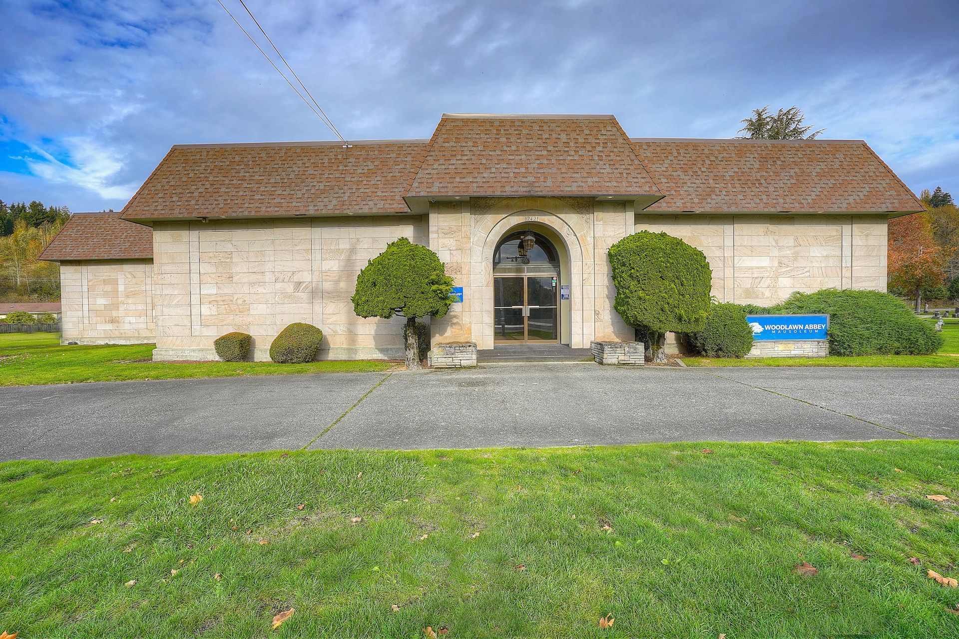 Stone house with arched entry, tan roof, gravel drive, and green lawn under a blue sky