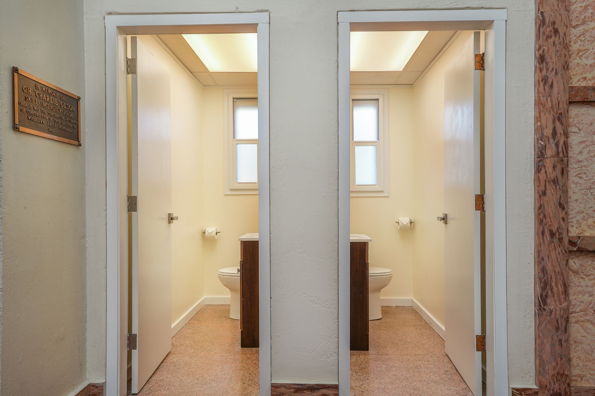 Two side-by-side bathroom stalls with open doors and beige walls in a narrow hallway.