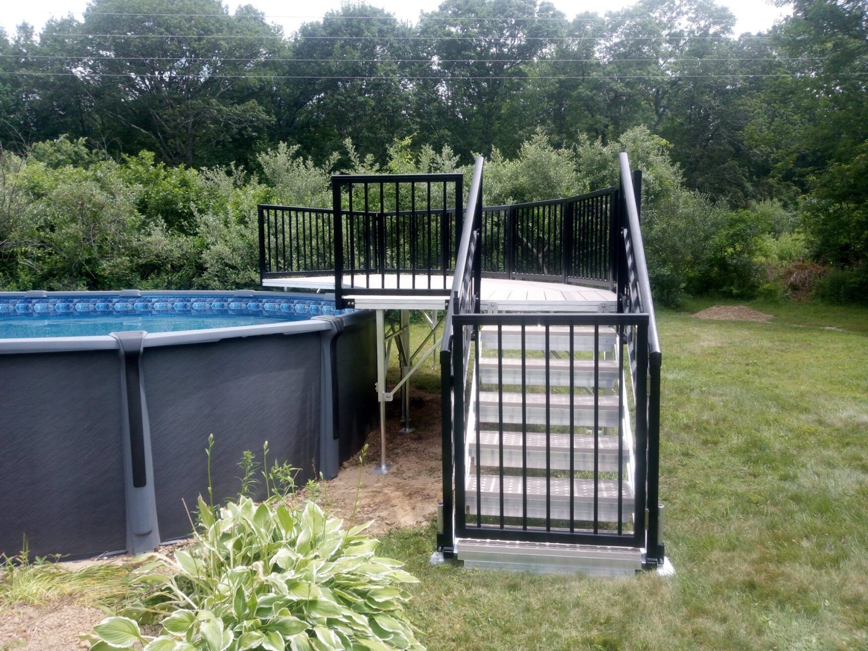 Backyard above-ground pool with black metal stairs and deck beside a grassy yard and trees
