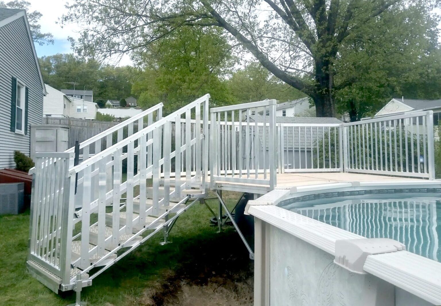 White wooden stairs and railing lead to a backyard deck beside an above-ground pool and house.