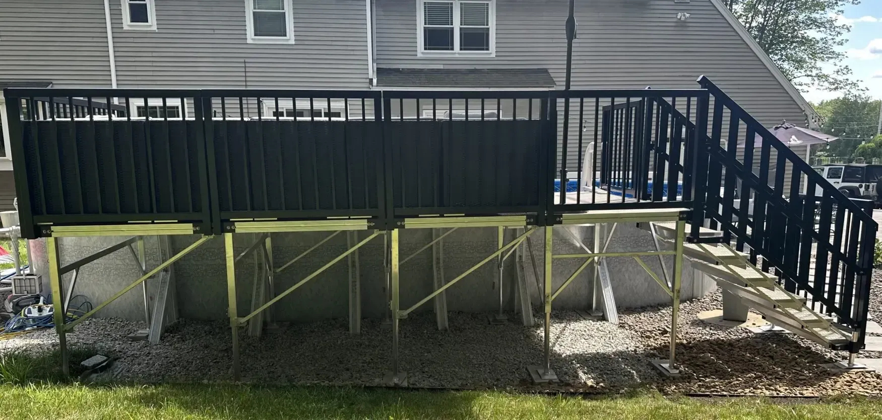 Raised wooden deck with black railing on support posts beside a house, with stairs on the right.