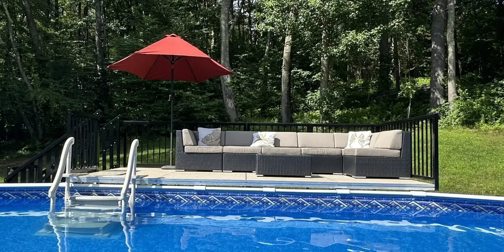 Backyard pool with blue water, white lounge chairs, gray seating area, and a red umbrella by trees