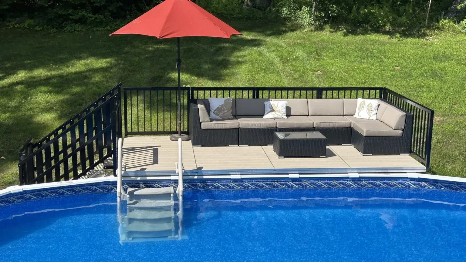 Backyard pool with deck, sectional sofa, red umbrella, and metal steps into the water