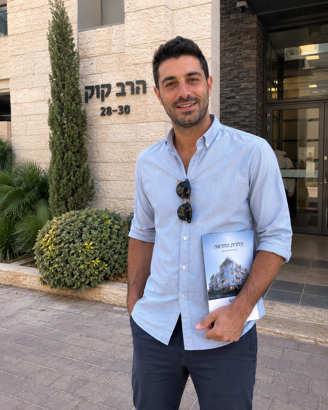 Man standing outside a building entrance, holding a brochure, wearing a light blue shirt and dark pants.