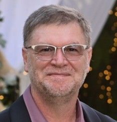 Close-up of a smiling man wearing glasses and a dark jacket at a softly lit event
