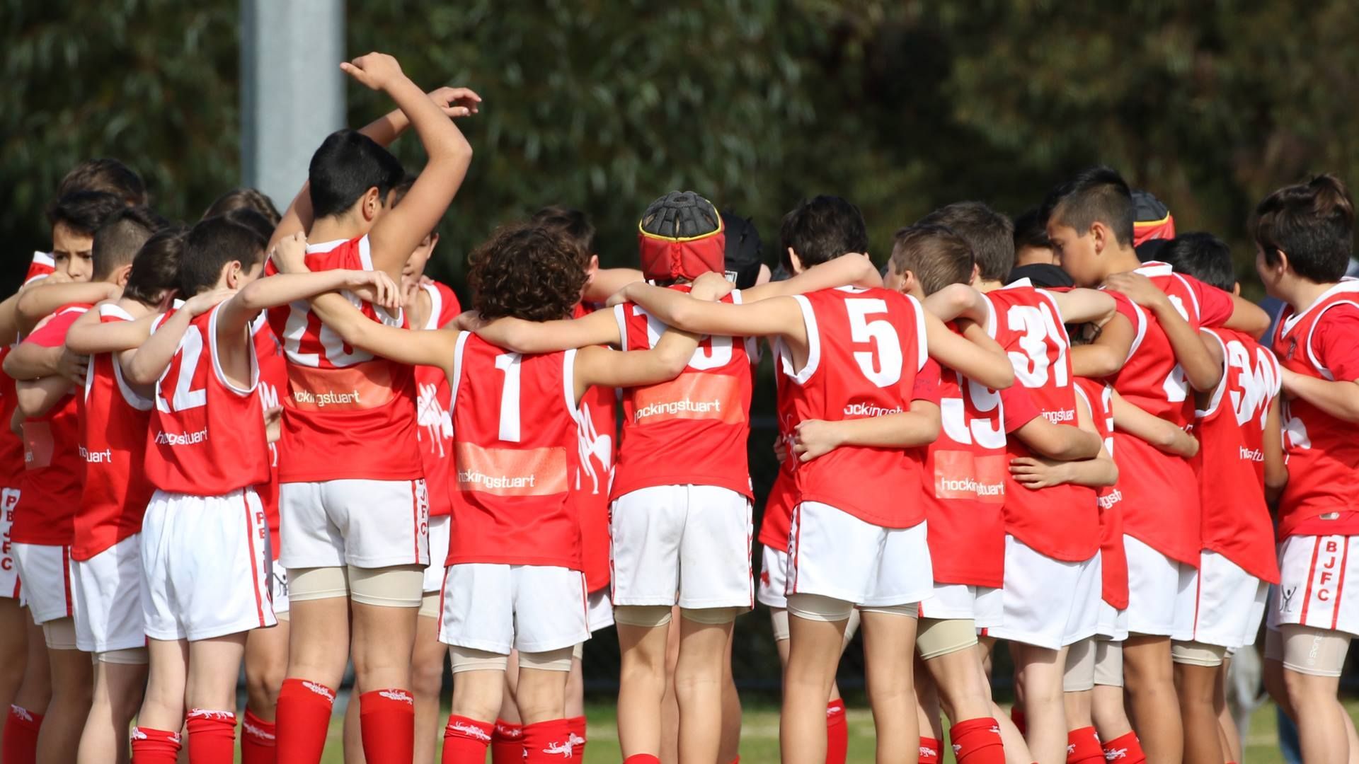 A group of team players in red and white uniforms huddle in a circle on a field, arms around each other's shoulders.