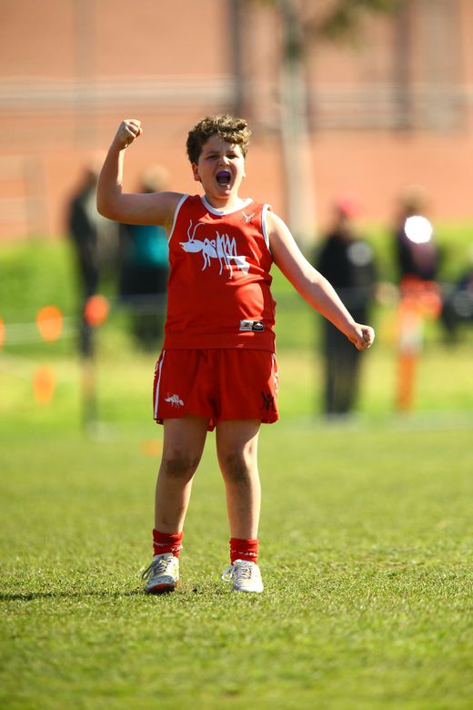 A child in a red uniform stands on a grass field, cheering with a raised fist and an excited expression.