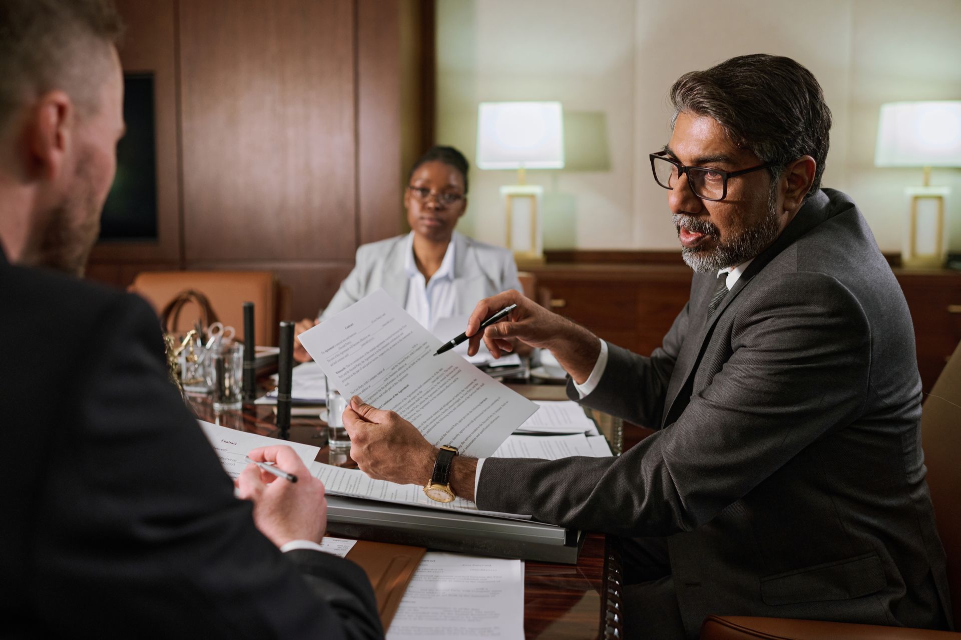 Three people in suits at a table. A man points to a document; another man sits opposite, and a woman sits behind them.