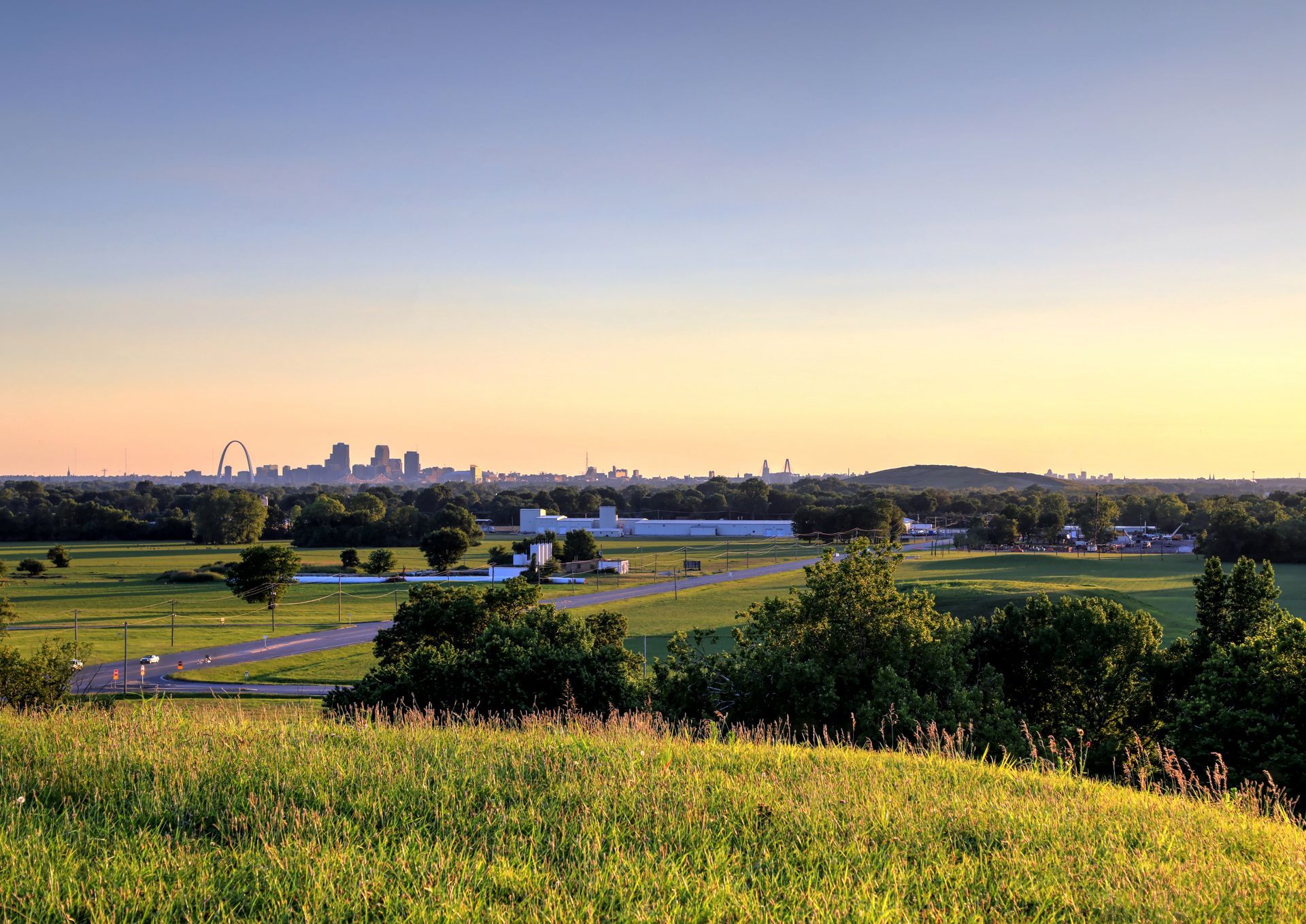 Green field with trees, buildings, and cityscape on the horizon under a light blue and yellow sky.