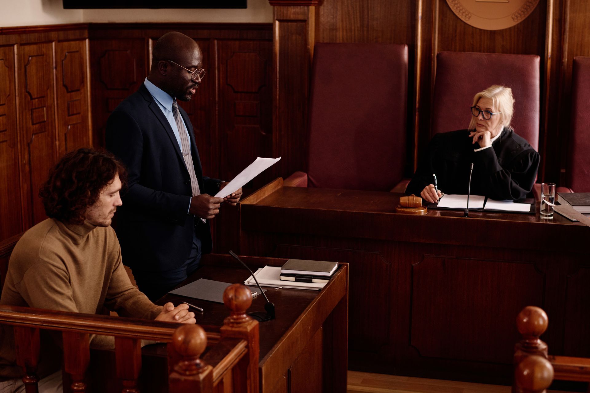 A lawyer speaks in court; a man sits at the defendant's table while a judge listens.