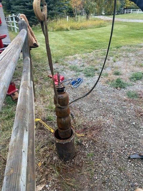 Rusted metal well pump standing in a grassy rural yard beside a wooden fence