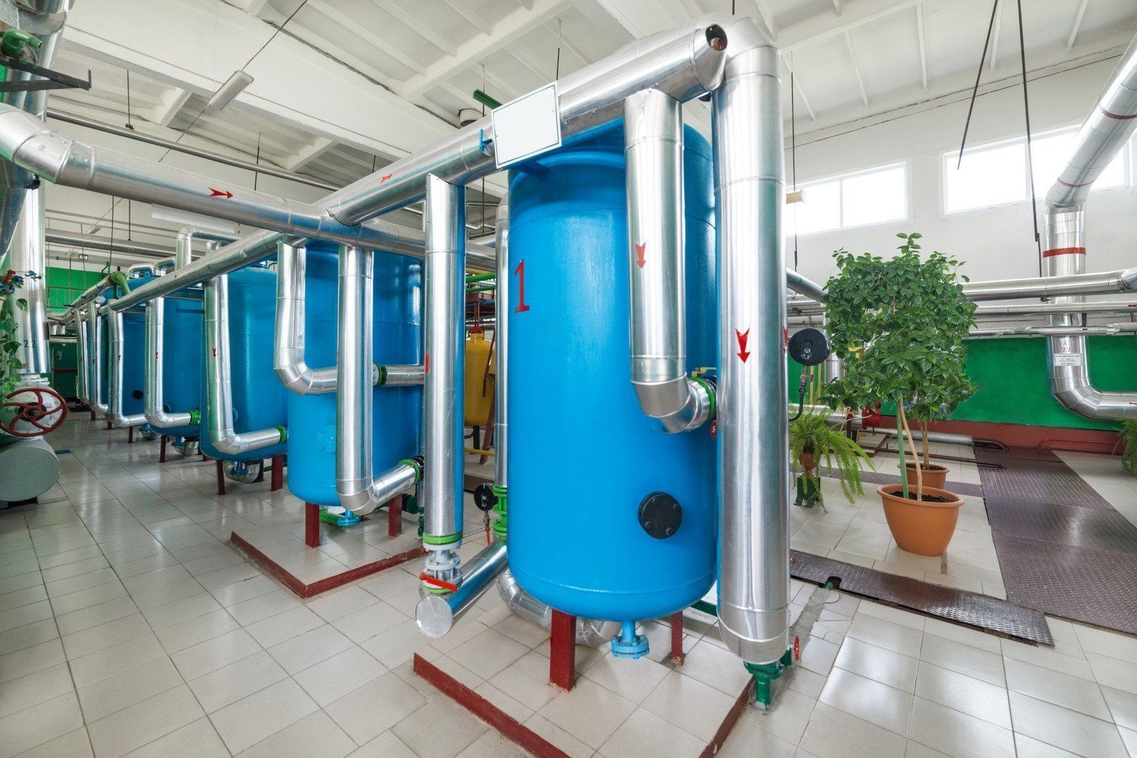 Blue industrial tanks and silver pipes in a clean plant room with tiled floor and potted plants