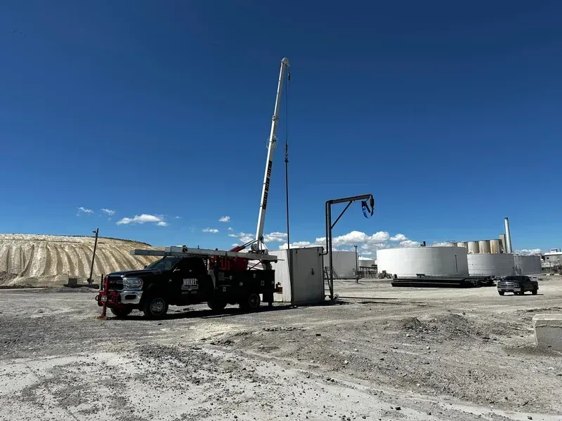 Truck with raised crane at an industrial site beside large white storage tanks under a clear blue sky