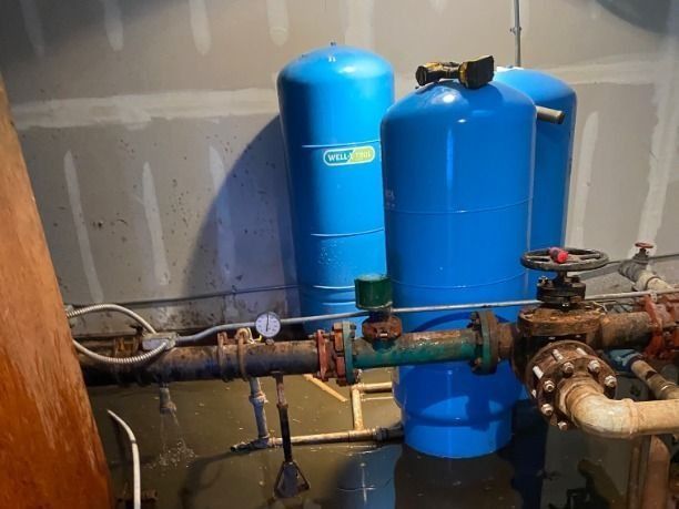 Industrial plumbing pipes with valves and gauges in a utility room, beside two blue water tanks