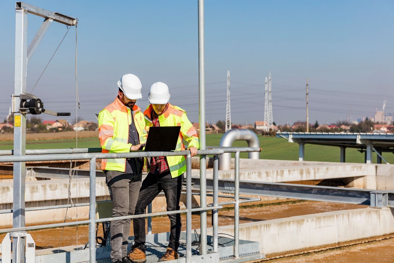 Two workers in hard hats reviewing a laptop at an industrial site with pipes and tanks