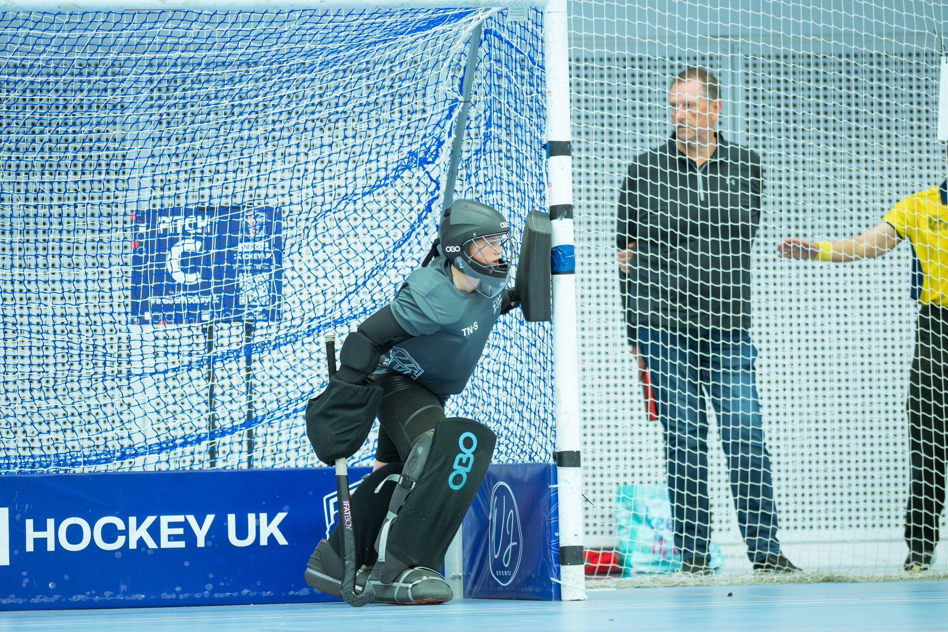 Hockey goalkeeper crouches by the net as a ball approaches, with spectators behind the goal.