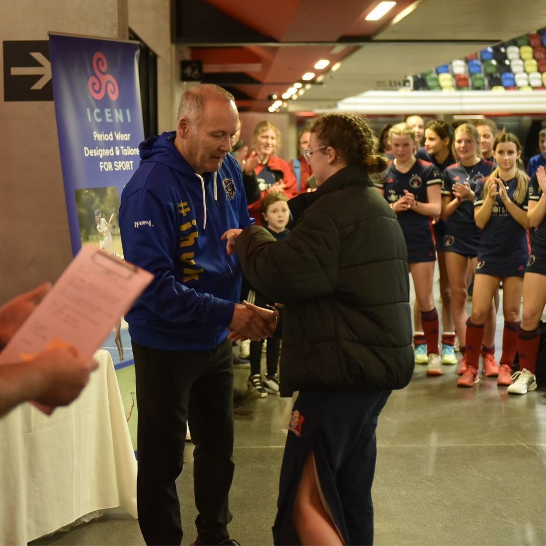 Presenter in blue jacket shakes hands with player while a team applauds behind them.