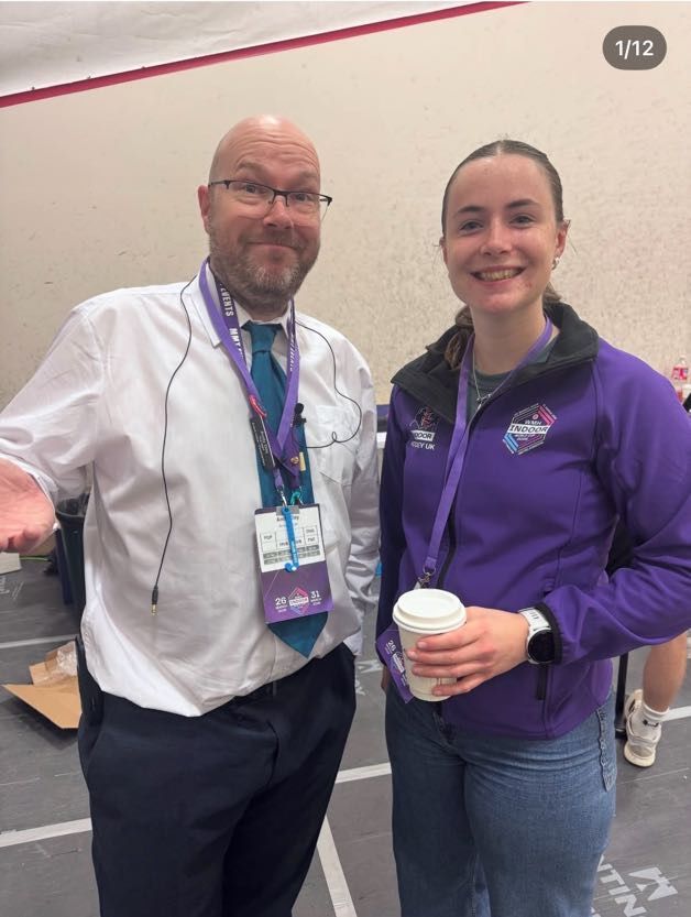 Two smiling volunteers stand indoors, one holding a coffee cup and the other wearing a lanyard