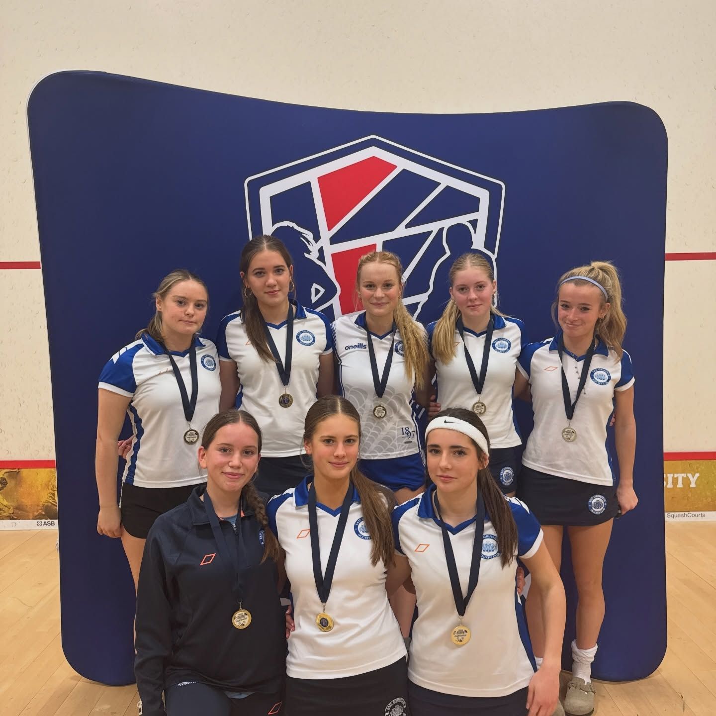 Six athletes in white jerseys pose with medals in front of a blue backdrop and sports logo.