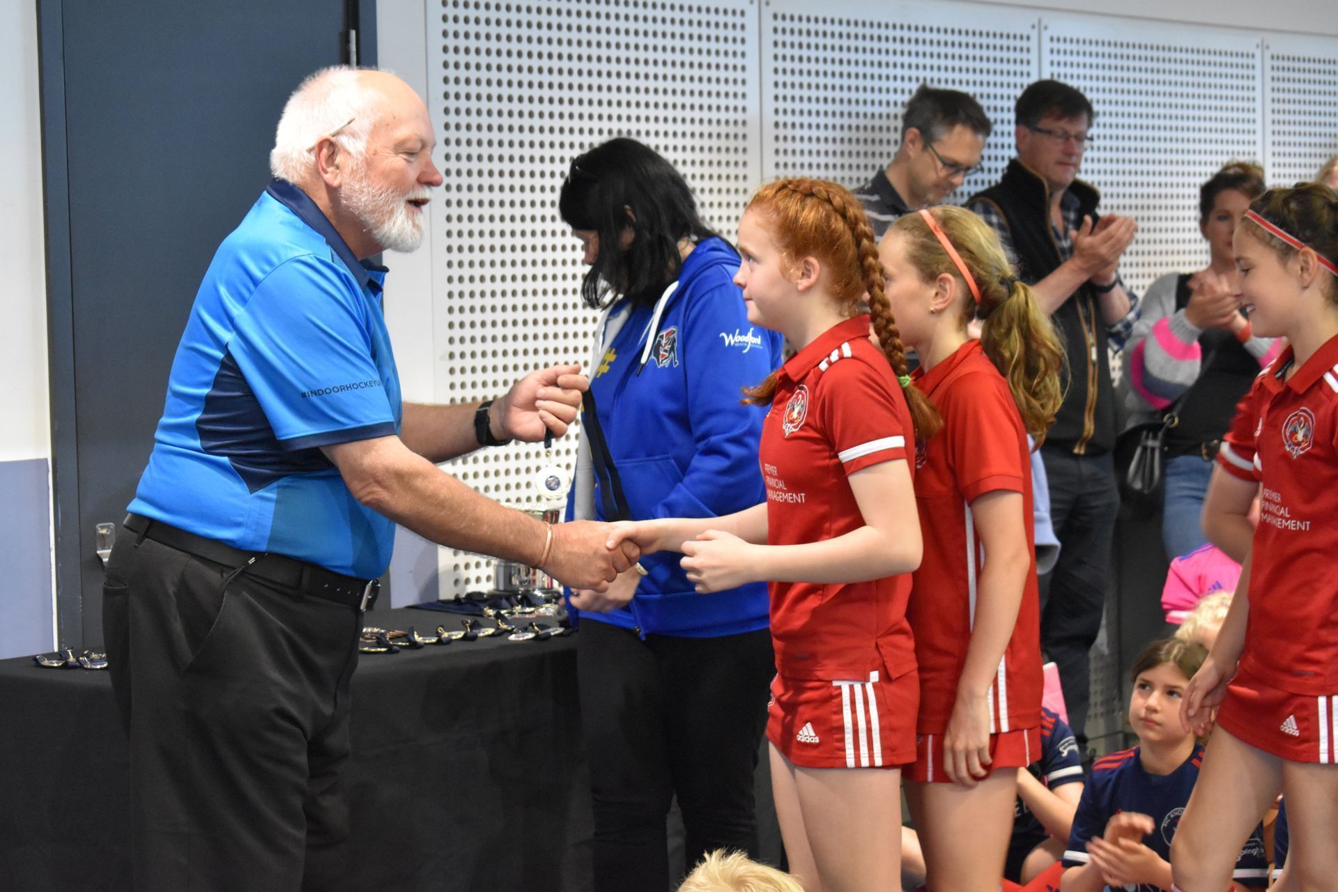 Presenter in blue shirt shaking hands with children in red sports uniforms indoors