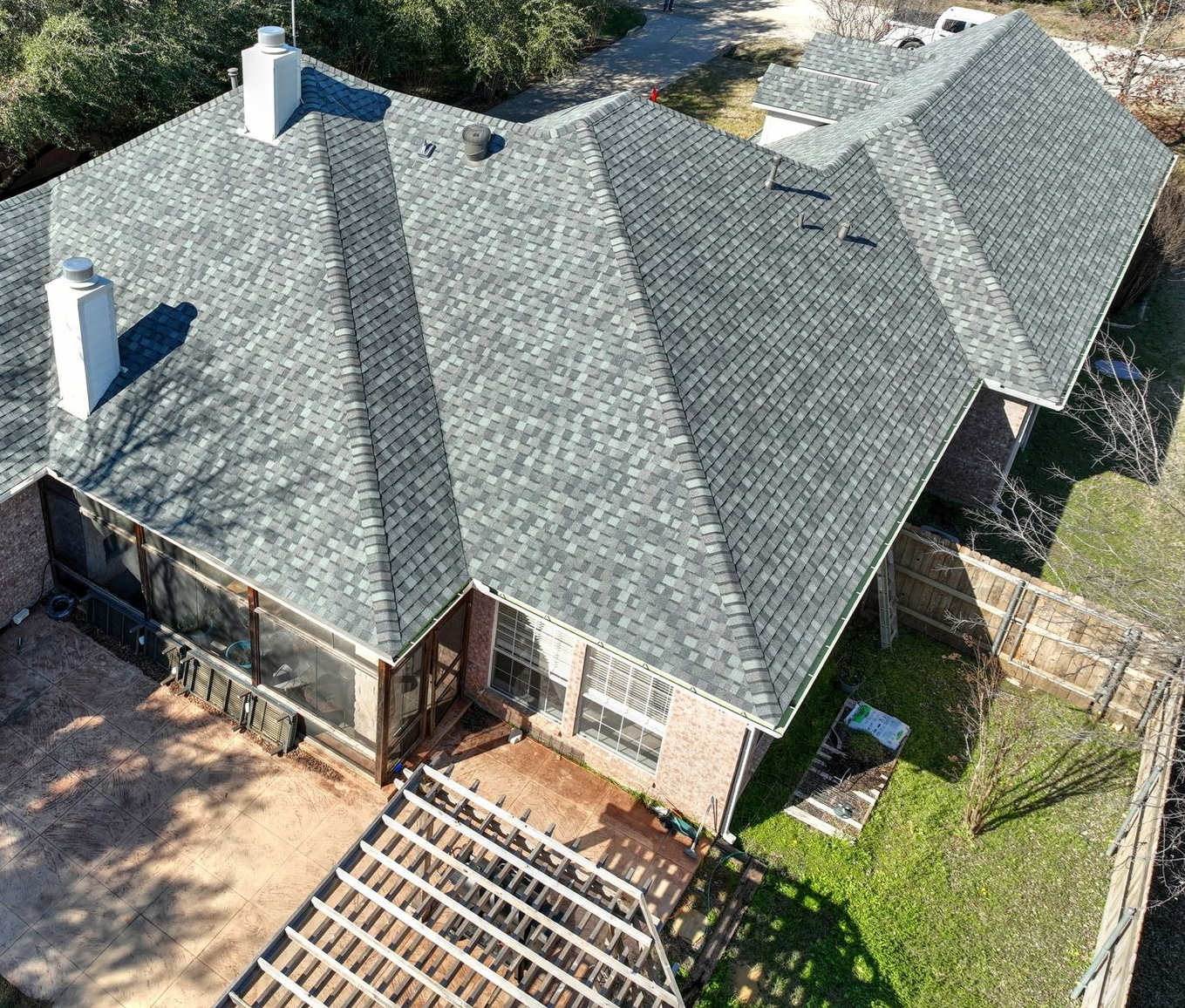 Aerial view of a brick house with a gray shingled roof, screened-in porch, and pergola on the patio.
