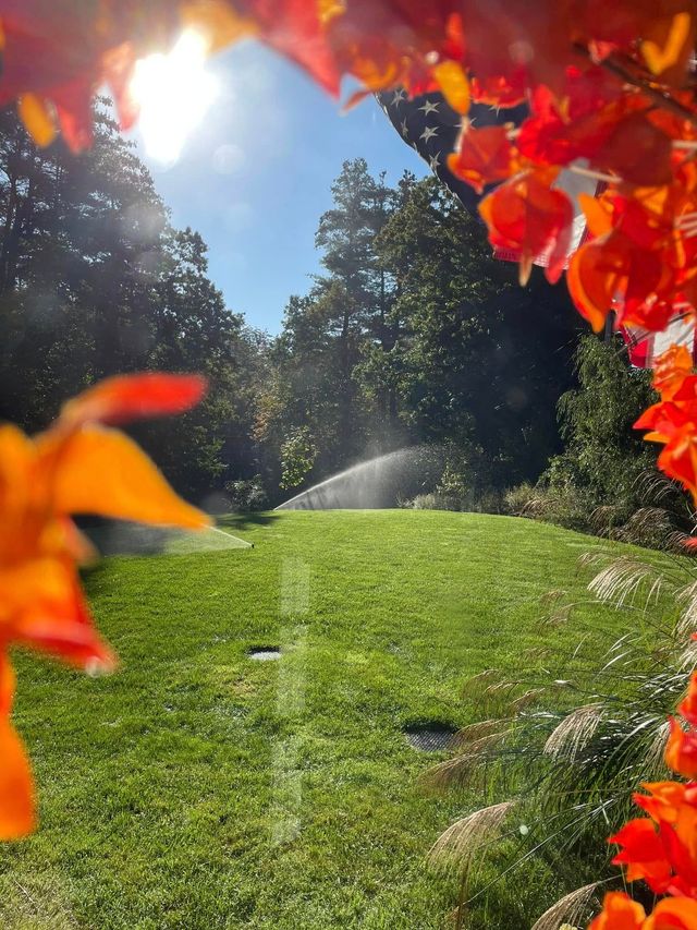 Sunlit green lawn with sprinklers, framed by bright red autumn leaves and trees